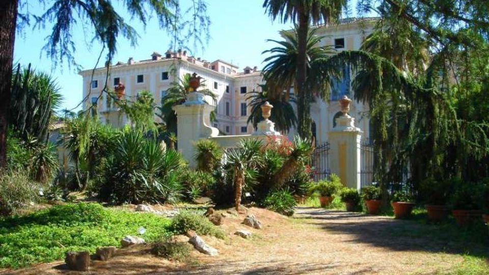 Viale sterrato in un giardino mediterraneo con palme, agavi e vegetazione fitta, che conduce a un cancello in ferro tra pilastri chiari con vasi ornamentali. Sullo sfondo si vede un grande edificio storico chiaro con più piani e finestre regolari.