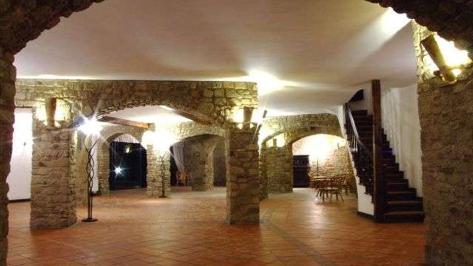 Ampio interno in stile rustico con pavimento in cotto e colonne in pietra che sorreggono una serie di archi, illuminati da appliques a parete. Sulla destra si vede una scala in legno che sale al piano superiore e, sullo sfondo, un’area con tavoli e sedie sotto le volte.