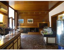 Interno di un bar con bancone lungo sulla sinistra, pareti e soffitto in legno e pavimento in marmo scuro. Sul fondo si vede un camino incassato e quadri alle pareti, mentre a destra ci sono una vetrina refrigerata e una vetrinetta espositiva.