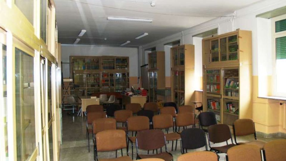 Aula interna con file di sedie imbottite disposte a platea, illuminata da lampade fluorescenti a soffitto. Lungo le pareti ci sono alte librerie e armadi vetrati con libri e materiali, con finestre sul lato destro.