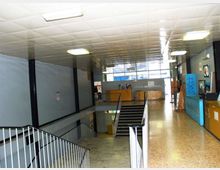 Interno di un edificio pubblico con ampio corridoio e pianerottolo che affaccia su una scala centrale, illuminato da plafoniere a soffitto. Pavimento in graniglia, pareti grigie con ringhiere metalliche e pannelli o bacheche lungo il lato destro.