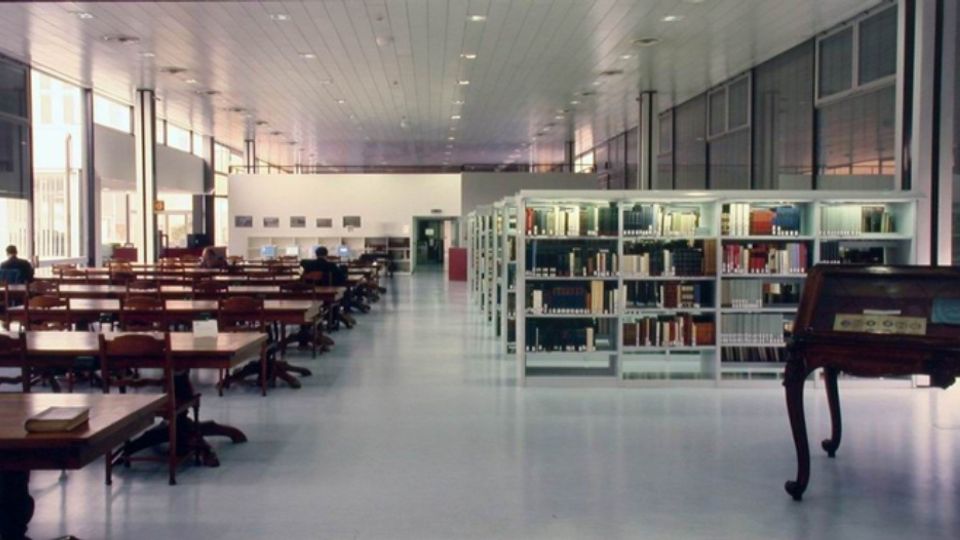 Ampia sala di biblioteca con lunghi tavoli e sedie in legno disposti in file, affiancati da scaffalature metalliche piene di libri. Ambiente moderno con soffitto alto a pannelli, pavimento chiaro e grandi finestre laterali che illuminano lo spazio.