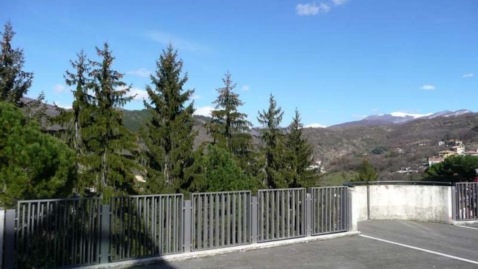 Terrazza o area pavimentata con ringhiera metallica lungo il bordo, affacciata su una valle con alberi sempreverdi e colline montuose. Sullo sfondo si vedono rilievi con chiazze di neve e un cielo azzurro con poche nuvole.