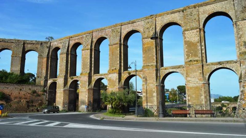 Grande acquedotto in pietra con alte arcate ripetute su due livelli, che attraversa una strada e si estende in diagonale sotto un cielo sereno. In primo piano si vedono carreggiata, segnaletica e una piccola area con lampione e panchine vicino alla base delle arcate.