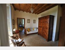 Interno di una camera in stile rustico con soffitto in legno a travi e lampadario, pavimento in cotto e pareti chiare. Arredamento con grande armadio in legno, divanetto con cuscini e tavolino vicino a una finestra, con porta aperta verso una stanza adiacente.