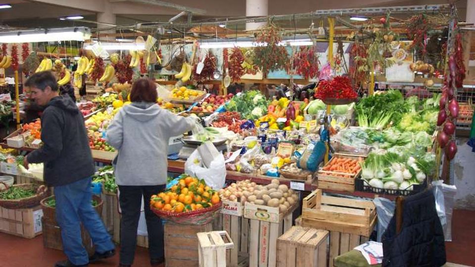 Interno di un mercato coperto con banco di frutta e verdura, cassette di legno e prodotti esposti su ampi tavoli. Dal soffitto pendono mazzi di peperoncini e banane, con scaffali e illuminazione a neon sullo sfondo.