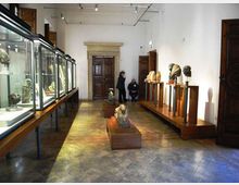 Interno di una sala museale con pareti bianche, pavimento in pietra e faretti a soffitto, con una grande porta in legno incorniciata da un portale in pietra sul fondo. Lungo la parete sinistra ci sono teche in vetro su supporti, mentre a destra e al centro sono esposte sculture e busti su piedistalli e basamenti in legno.