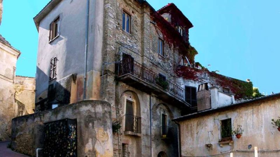 Edificio storico in pietra e intonaco su una strada in salita, con balconi in ferro battuto e finestre irregolari. Alla base si vedono muri di contenimento e un passaggio ad arco, con vegetazione che ricopre parzialmente il tetto e la facciata.
