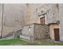 Cortile in pietra con scala laterale e muretto in muratura, addossati a una parete di edificio in pietra e intonaco decorato. Si vedono due porte in legno con cornici in pietra e un piccolo stemma sulla parete.
