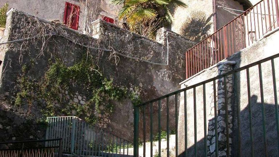 Scalinata in pietra in un borgo collinare, con ringhiere metalliche e muri in pietra ricoperti in parte da vegetazione. In alto si vedono case intonacate con persiane rosse e una palma sotto un cielo sereno.