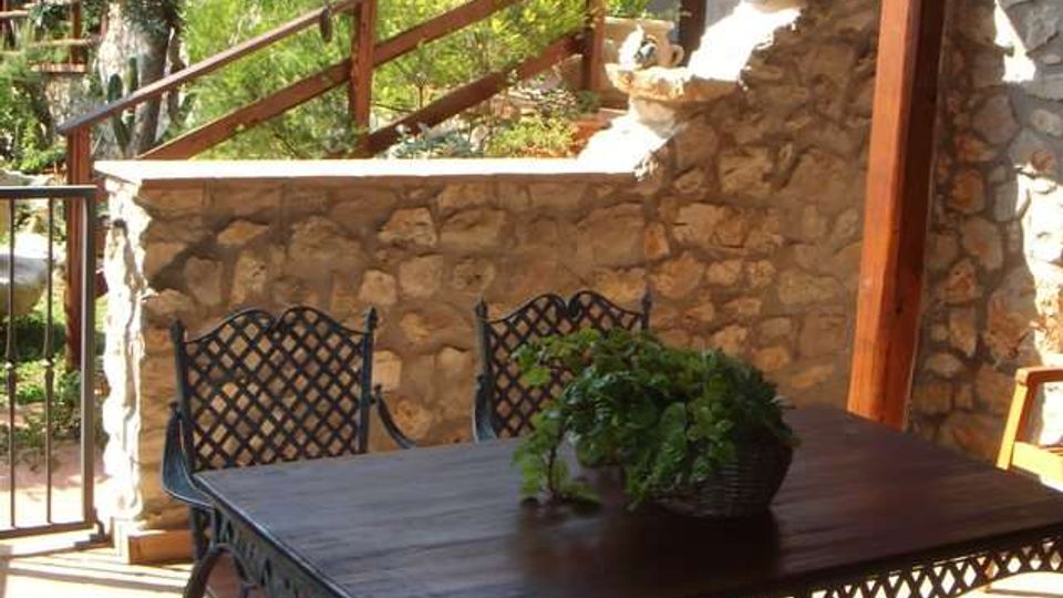 Terrazza coperta con travi in legno, arredata con tavolo e sedie in ferro battuto su pavimento in cotto. Sullo sfondo si vede un muretto in pietra con vegetazione e una costruzione con pareti in pietra e grandi finestre affacciate sul giardino.