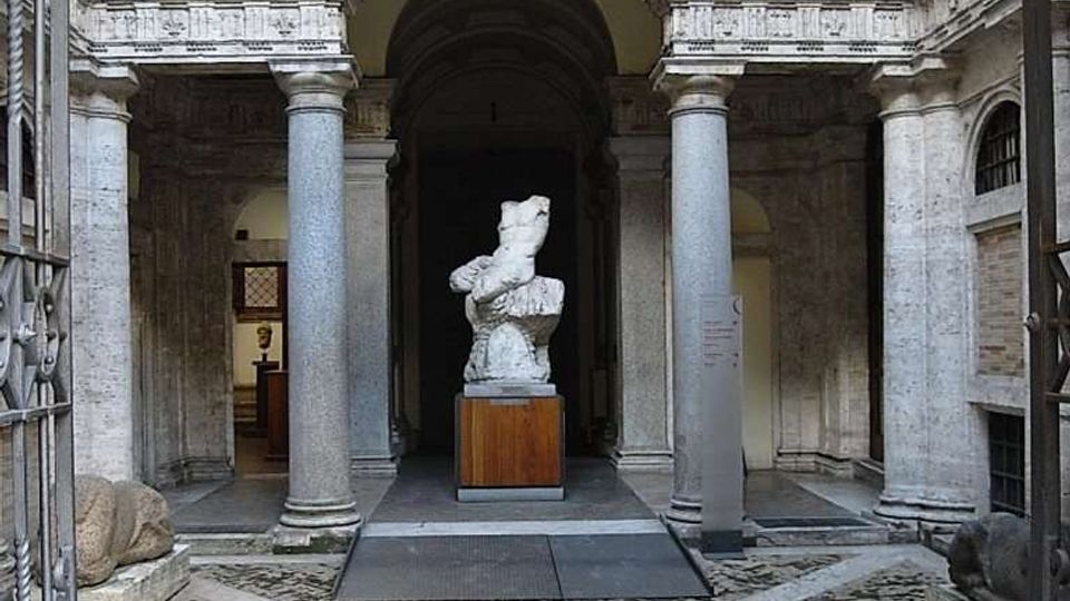 Cortile interno di un edificio storico con pavimentazione in pietra a motivi geometrici, colonne in pietra e un grande arco centrale che introduce a un portico voltato. Al centro è esposta una statua su basamento, circondata da pareti in laterizio e intonaco con aperture laterali.