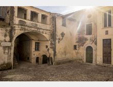 Piccola piazza lastricata in pietra circondata da edifici storici in intonaco consumato, con un passaggio ad arco e loggiato superiore, lanterna a muro e portali in legno. La luce del sole entra dall’angolo in alto a destra, evidenziando le facciate con finestre e persiane.