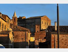 Vista di un borgo storico con case in pietra dai tetti in tegole, vicoli stretti e un campanile con guglia sullo sfondo. La luce calda del tramonto illumina le facciate, con lampioni e antenne visibili tra gli edifici.