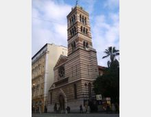 Chiesa in stile romanico-gotico con facciata a fasce orizzontali bianche e rosse e alto campanile con logge ad archi sovrapposti. L’edificio si affaccia su una piazza urbana, con palazzi adiacenti e una palma sul lato destro.
