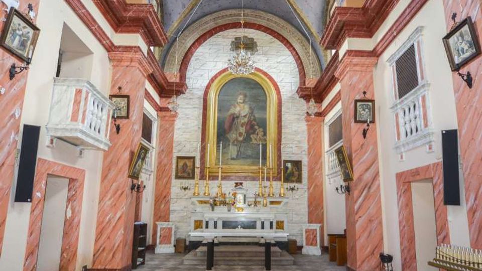 Interno di una chiesa con navata centrale che conduce a un altare in marmo bianco, sovrastato da una grande pala dipinta entro un’arca dorata e un lampadario. Le pareti laterali hanno colonne e cornici in marmo rosato con balconate bianche, mentre il soffitto è a volta decorata con motivi dipinti.