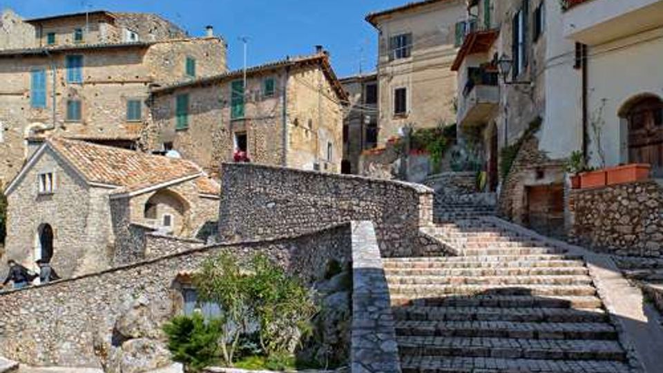 Scalinata in pietra che sale tra case in muratura con facciate chiare, persiane e balconi, in un borgo storico collinare. Muri e pavimentazioni in pietra delimitano piccoli terrazzamenti con vegetazione sotto un cielo sereno.