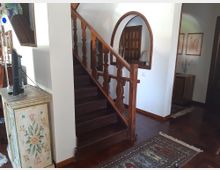 Interno di una casa con scala in legno scuro e corrimano intagliato che sale lungo una parete bianca. Sul pianerottolo si vede una grande specchiera ad arco, pavimento in parquet con tappeto e mobili in legno, tra cui una cassettiera dipinta e una piccola zona con quadri alle pareti.