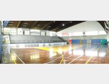 Interno di un palazzetto dello sport con campo polivalente in parquet lucido e linee tracciate per diversi sport. Sul lato lungo si vedono ampie gradinate, pareti con pannelli colorati e un soffitto alto con travi e illuminazione.