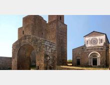 Complesso medievale in pietra con grande arco d’ingresso e massicce torri quadrangolari, accanto a una chiesa romanica con facciata a capanna, rosone centrale e portale ad arco. L’insieme è in un’area aperta con terreno erboso e cielo sereno sullo sfondo.