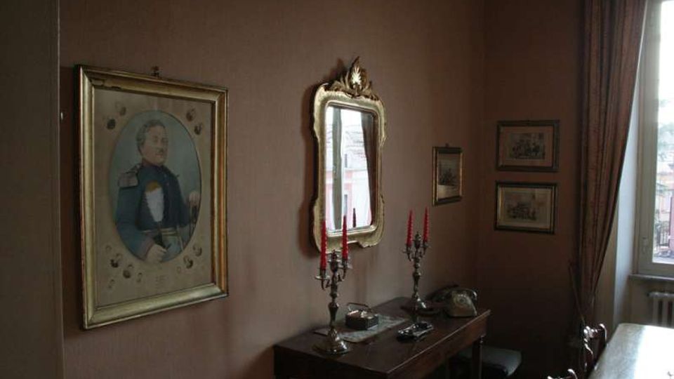 Interno di una stanza in stile classico con pareti marroni, un tavolo consolle in legno con candelabri e oggetti decorativi, e uno specchio con cornice dorata sopra. Sulle pareti sono appesi un ritratto incorniciato e alcune stampe, accanto a una finestra con tende pesanti.