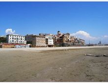 Ampia spiaggia sabbiosa in primo piano con lungomare e fila di edifici storici sullo sfondo, tra cui una torre cilindrica fortificata vicino alla costa. Cielo sereno e, sulla destra, area portuale con alberi di barche visibili in lontananza.