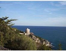 Torre costiera in pietra su un promontorio roccioso, circondata da vegetazione mediterranea, con il mare aperto sullo sfondo sotto un cielo sereno.