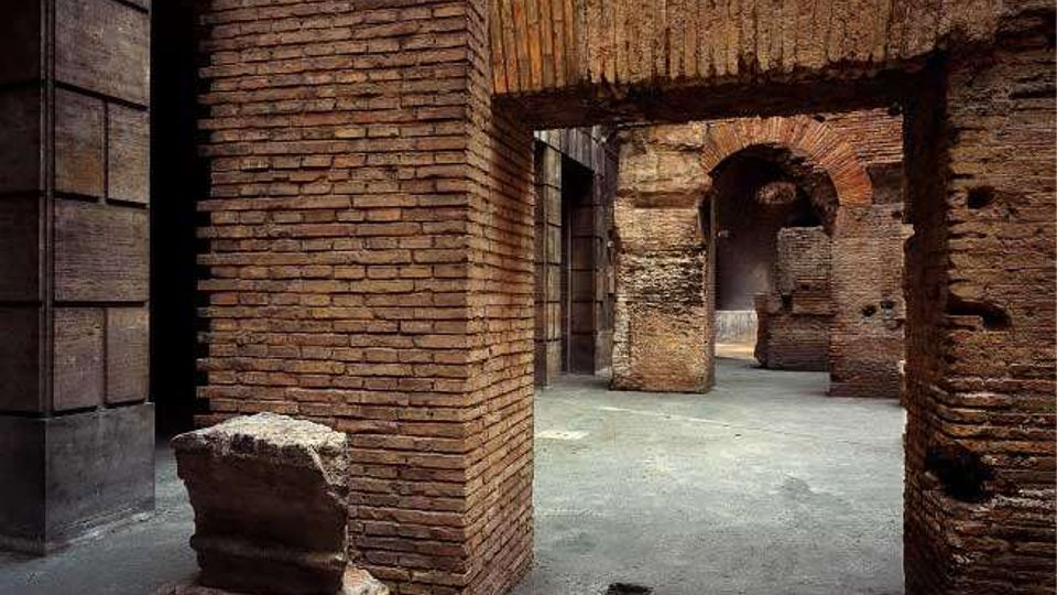 Interno di rovine in mattoni con grandi archi e passaggi, con pareti antiche segnate da fori e aperture. Pavimento in cemento e un blocco di pietra spezzato in primo piano, affiancati da una parete moderna rivestita in lastre scure.