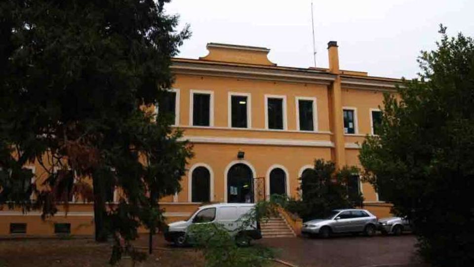 Edificio a due piani color ocra con finestre rettangolari al piano superiore e ingressi ad arco al piano terra, con una breve scalinata centrale. Davanti si vede un vialetto con auto parcheggiate e un’area con alberi e vegetazione che incorniciano la facciata.