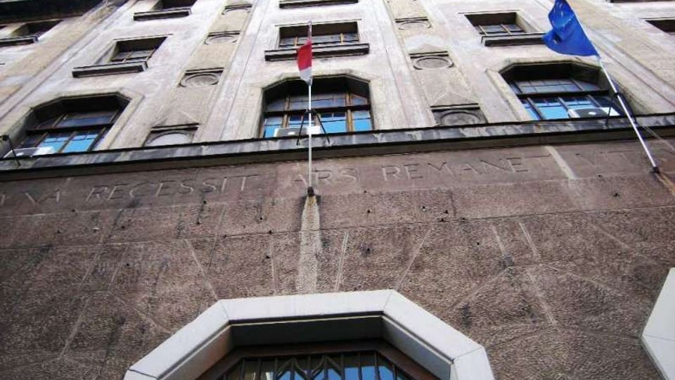 Facciata di un edificio istituzionale in pietra vista dal basso, con più piani di finestre rettangolari e un grande portale ad arco con inferriate. Due aste portabandiera sulla parete espongono bandiere, e una scritta incisa corre lungo la fascia sopra l’ingresso.