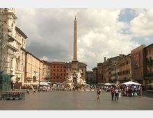 Ampia piazza urbana circondata da palazzi storici, con al centro una fontana monumentale con sculture e un alto obelisco. Ai lati della piazza si vedono edifici color ocra e arancio, aree con tende e bancarelle, sotto un cielo parzialmente nuvoloso.