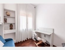 Piccolo studio domestico con pareti bianche, pavimento in legno scuro e finestra con tende leggere. A destra c’è una scrivania bianca con cassettiera e sedia ergonomica, mentre a sinistra si vedono mensole a incasso e un divano blu.