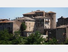 Complesso di edifici storici in pietra con tetti in coppi, collegati tra loro e affiancati da una torre cilindrica tipo fortilizio. In primo piano ci sono alberi e vegetazione, con un piccolo passaggio ad arco tra le costruzioni sotto un cielo limpido.