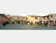 Piazza lastricata in pietra in un borgo storico, circondata da case in muratura dai toni ocra e rosa con persiane, balconi e vasi di piante. Sullo sfondo si vede un arco in pietra inserito nelle mura, che conduce a una strada oltre il centro abitato.