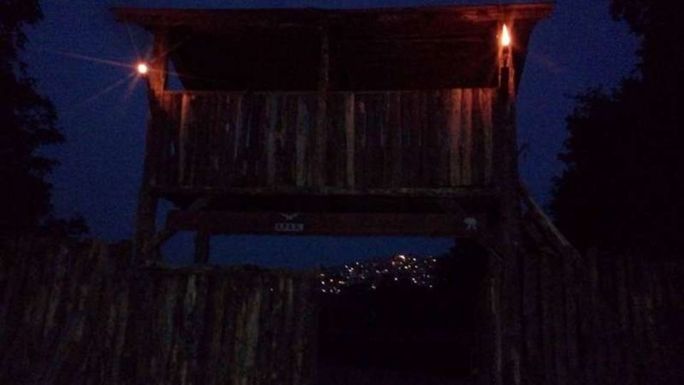 Capanna sopraelevata in legno con parapetto e tetto sporgente, illuminata da due fiaccole ai lati in un’ambientazione notturna. In basso si vedono una staccionata di tronchi e luci lontane sullo sfondo, con alberi ai margini della scena.