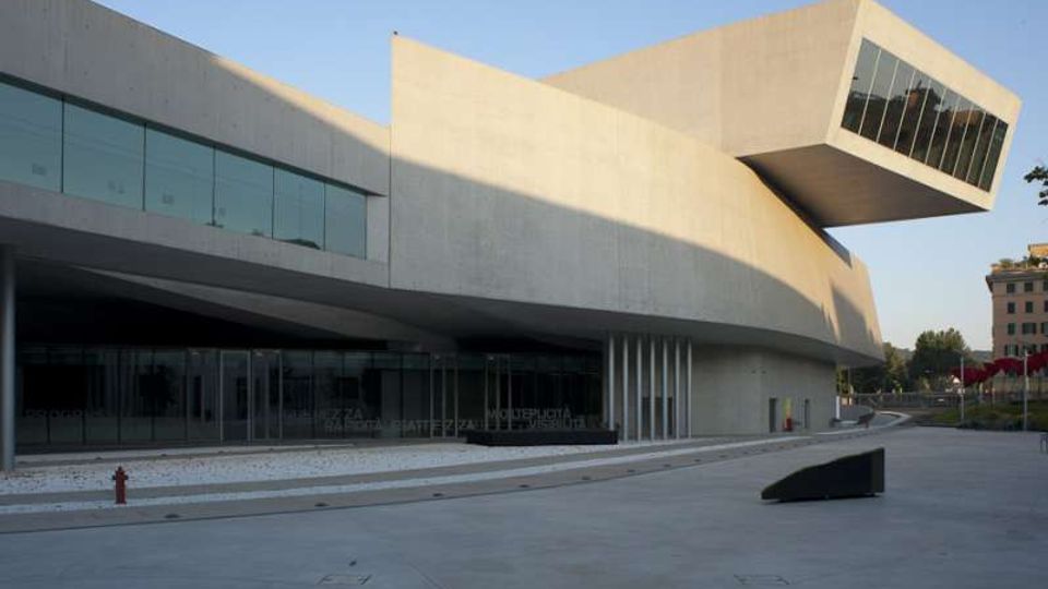 Edificio moderno in cemento e vetro con volumi curvilinei e un grande corpo sporgente vetrato a sbalzo. Davanti si estende un’ampia piazza pavimentata con ingresso vetrato e pochi elementi urbani.