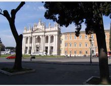 Facciata di un grande edificio storico in stile neoclassico con portico a colonne, frontone e statue sul tetto, affacciato su una piazza ampia. In primo piano si vedono alberi che incorniciano la scena e una strada con lampioni e alcune auto parcheggiate.