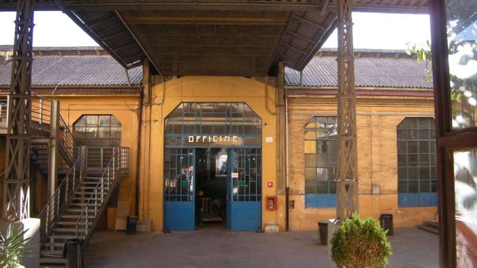 Cortile interno di un complesso industriale con tettoia in lamiera sostenuta da pilastri metallici e una grande porta vetrata blu con insegna “OFFICINE”. Ai lati si vedono finestre ad arco, una scala metallica esterna e muri in mattoni gialli.
