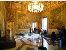 Sala storica con pareti interamente decorate da grandi affreschi e cornici dorate, soffitto ornato e grande lampadario centrale. Pavimento geometrico in marmo e alte finestre laterali, con alcuni elementi e arredi coperti o in allestimento.
