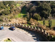 Parcheggio asfaltato curvo affacciato su una valle verde, delimitato da un muretto in pietra con merlature. Sullo sfondo si vede un pendio boscoso con alberi e cespugli e un’area di prato che si apre tra la vegetazione.