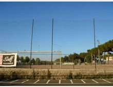 Campo sportivo all’aperto recintato con rete alta e pali metallici, visto da un parcheggio con strisce bianche. Sullo sfondo si notano alberi, un palo faro e un cartellone pubblicitario sul lato sinistro sotto un cielo sereno.