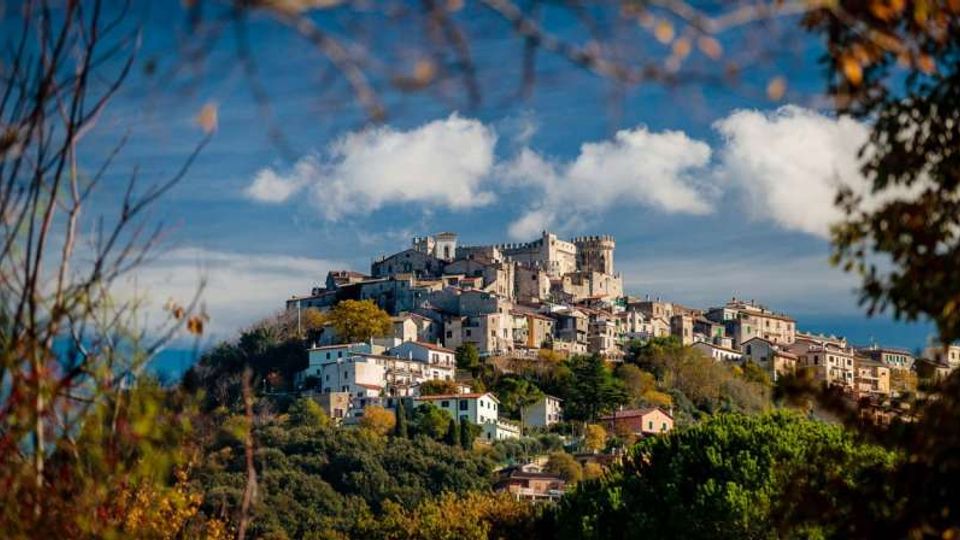 Borgo collinare con case in pietra e tetti chiari disposte a terrazze, dominato da un grande castello-fortezza con torri e mura sulla sommità. In primo piano vegetazione e alberi, con cielo azzurro e nuvole sullo sfondo.