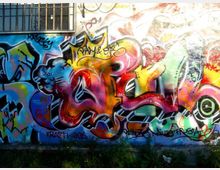 Muro esterno di un edificio ricoperto da un grande murale di graffiti multicolori con forme astratte e scritte sovrapposte. In alto è visibile una finestra con inferriate, mentre alla base del muro ci sono erba e vegetazione.