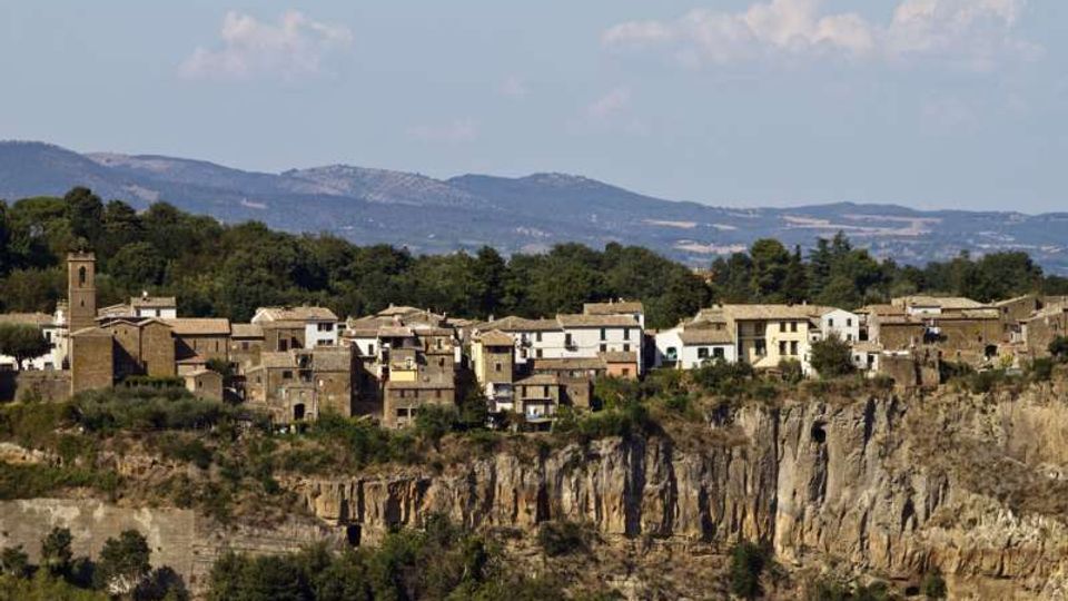 Panorama di un borgo storico con case in pietra e una torre campanaria, disposto lungo il bordo di una scogliera rocciosa. Sullo sfondo si vedono colline e montagne con boschi sotto un cielo parzialmente nuvoloso.