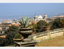 Vista panoramica da una terrazza in pietra con vaso ornamentale e pianta grassa in primo piano, affiancata da altri vasi lungo un parapetto. Sullo sfondo si vedono tetti in terracotta e una chiesa con cupola e campanili, con la città e la pianura in lontananza.