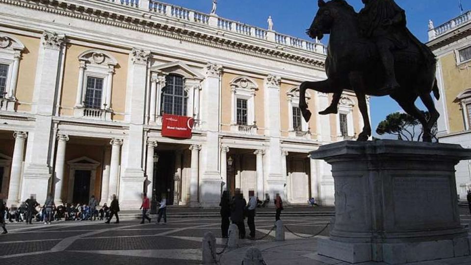 Piazza con grande statua equestre in bronzo su alto basamento di pietra, in primo piano. Sullo sfondo si vede un palazzo storico in stile classico con facciata chiara, portico ad archi, finestre ornate e balaustra con statue sul tetto.