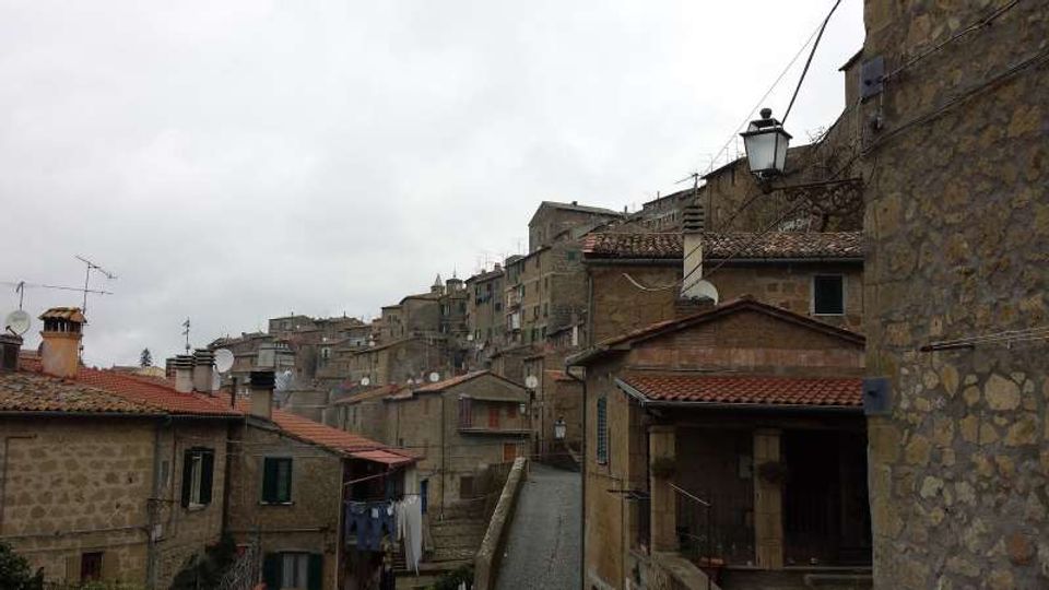 Veduta di un borgo collinare con case in pietra e tetti in coppi rossi, disposte su pendii e vicoli stretti. In primo piano si vede una strada in salita tra edifici, con lampione, antenne e camini sotto un cielo nuvoloso.