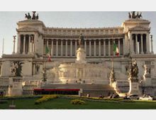 Veduta frontale del Vittoriano (Altare della Patria) a Roma, grande monumento in marmo bianco con lunga fila di colonne, ampie scalinate e statue equestri ai lati. Davanti si vede un prato curato con aiuole, bandiere italiane e una strada con veicoli.