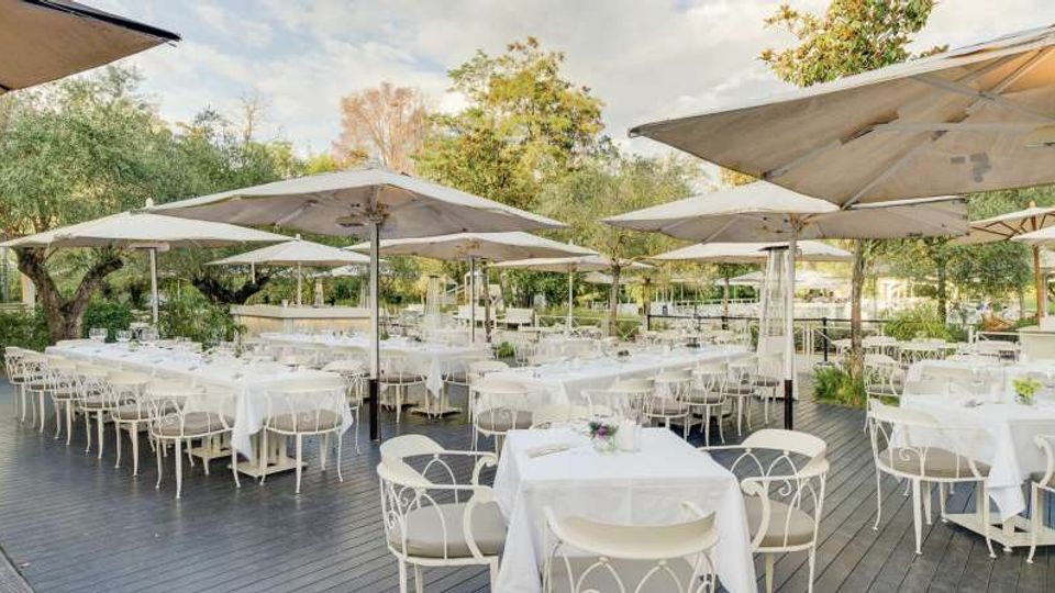 Terrazza all’aperto di un ristorante con tavoli apparecchiati con tovaglie bianche e sedie in metallo, disposti su una pedana in legno. Grandi ombrelloni chiari coprono l’area, circondata da alberi e vegetazione.
