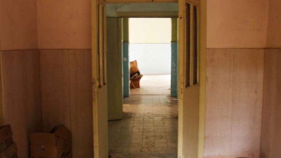 Interno di un edificio spoglio con pavimento in graniglia e pareti intonacate, con una doppia porta a battente aperta al centro. Attraverso l’ingresso si vede un corridoio con rivestimento a parete e luce naturale proveniente da un’apertura sul fondo.